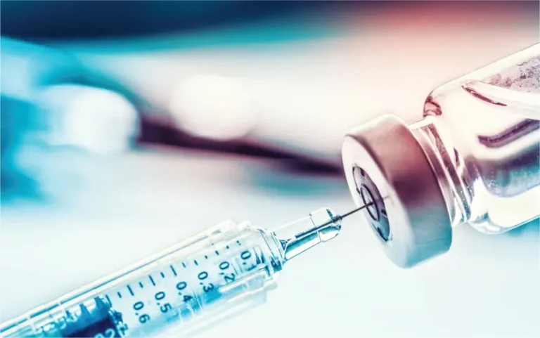 A close-up of a syringe needle inserted into the rubber stopper of a glass medicine vial, with a blurred medical background.