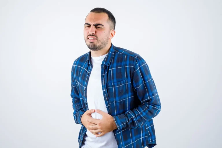 A man in a blue plaid shirt and white t-shirt stands against a plain background, holding his stomach with both hands and wincing in pain, suggesting he has a stomachache.