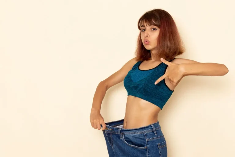 A young woman in a teal sports bra shows off her weight loss by wearing oversized jeans and pulling the waistband away from her waist, making a playful face against a plain beige background.