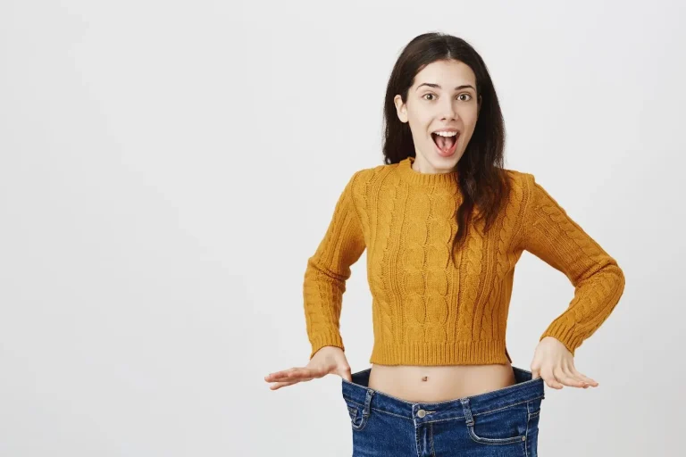A surprised woman in a yellow sweater holds out the waistband of oversized jeans, revealing how loose they are against her waist, standing in front of a plain white background.