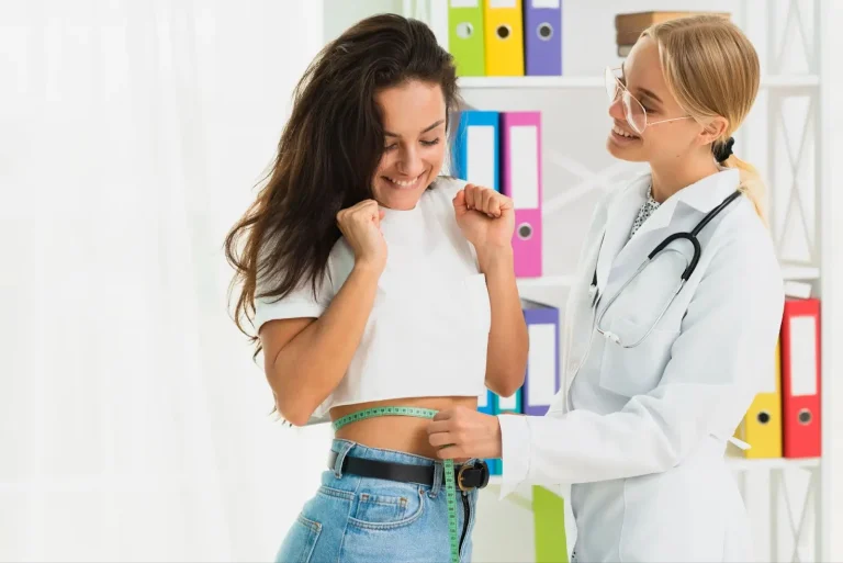 A doctor measures a smiling woman's waist with a tape measure in a bright office. The woman looks happy and excited, while the doctor smiles and wears a white coat and stethoscope. Colorful binders are visible in the background.