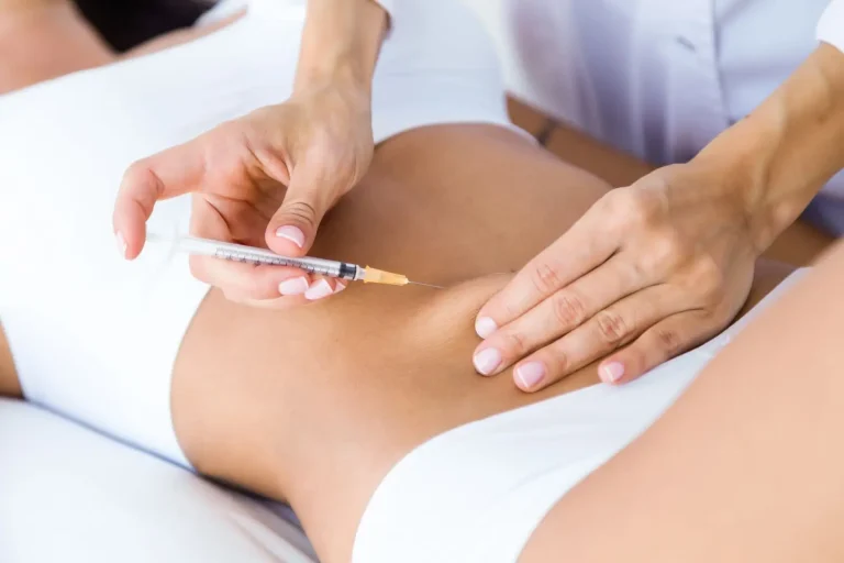 A person receiving an injection in the abdomen from a medical professional, who is wearing a white lab coat. The patient is lying down and wearing white clothing.