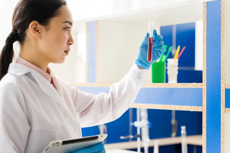 A scientist wearing a white lab coat and blue gloves examines a test tube containing a red liquid in a laboratory, holding a tablet in her other hand.