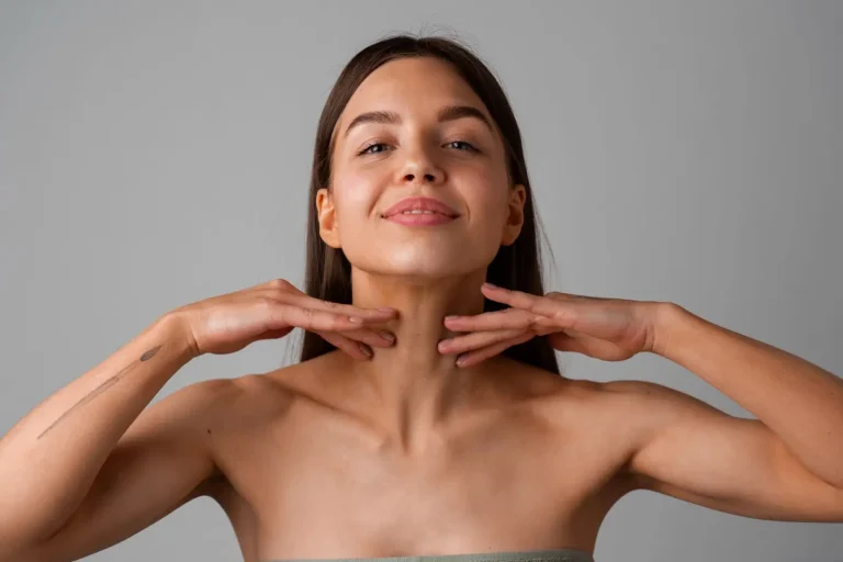 A woman with long brown hair and bare shoulders smiles slightly while gently touching her neck with both hands, set against a plain gray background.