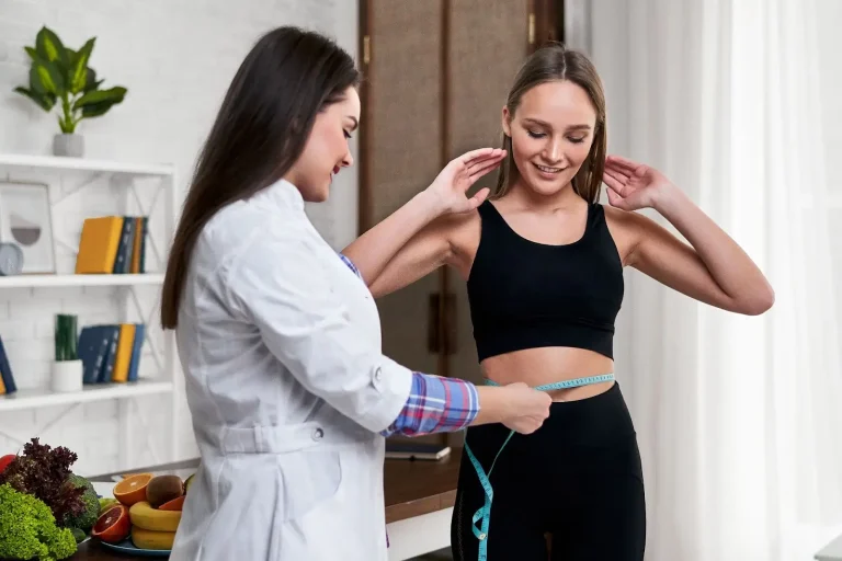 A woman in fitness clothing smiles as a nutritionist or doctor measures her waist with a tape measure in a bright room with fruits and vegetables on the table nearby.