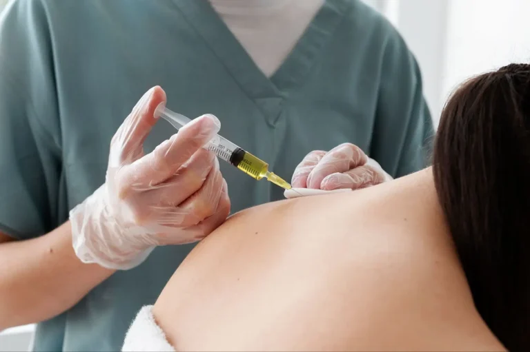 A healthcare professional in scrubs and gloves injects a yellowish liquid into the upper back of a patient with bare shoulders, suggesting a medical or therapeutic procedure.