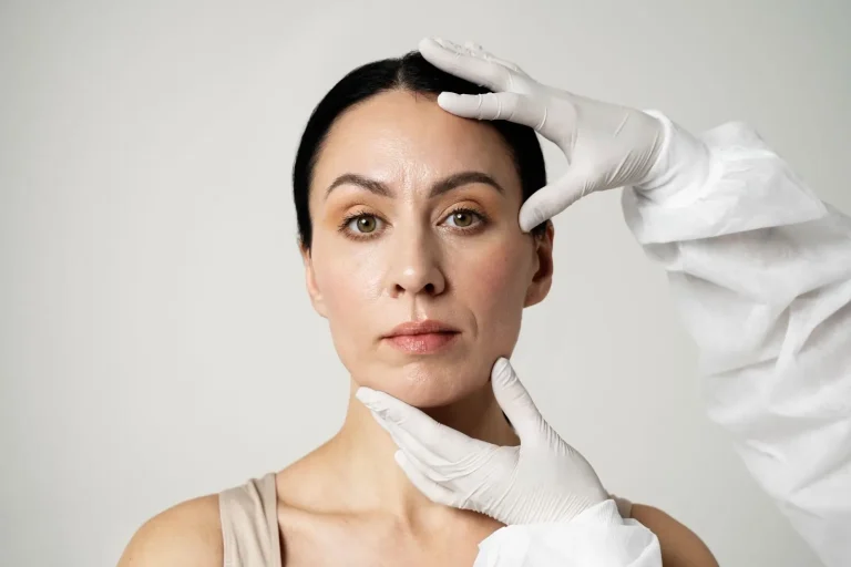 A woman with dark hair is examined by a person wearing white gloves and a white coat, with gloved hands gently touching her forehead and chin. The background is plain and light-colored.