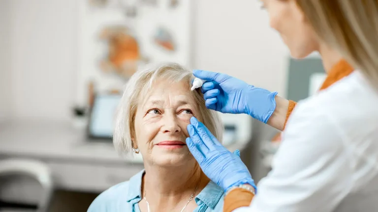 A healthcare professional wearing blue gloves applies eye drops to an older woman who is seated and looking up, in a medical office setting.