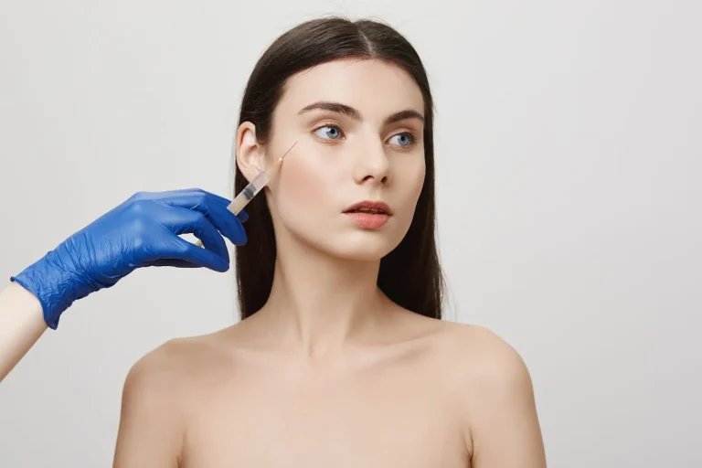 A woman with long dark hair and bare shoulders looks to the side while a gloved hand holds a syringe near her cheek, suggesting a cosmetic or dermatological procedure. The background is plain and light-colored.