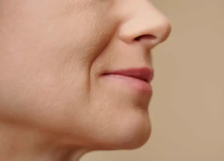 Close-up of the lower half of a person's face, showing smooth skin, slight smile lines, and natural lips against a neutral beige background.