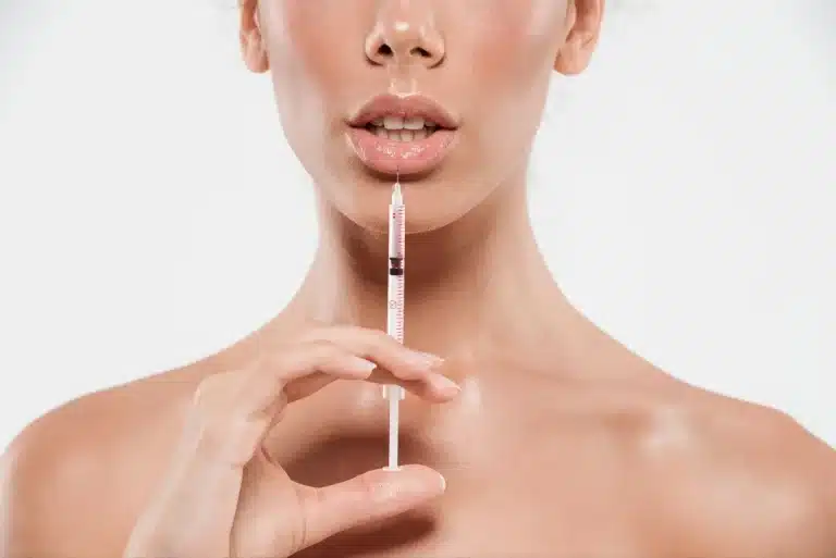 A woman holds a syringe upright near her slightly parted lips, suggesting an impending cosmetic procedure such as lip injections, against a plain white background.