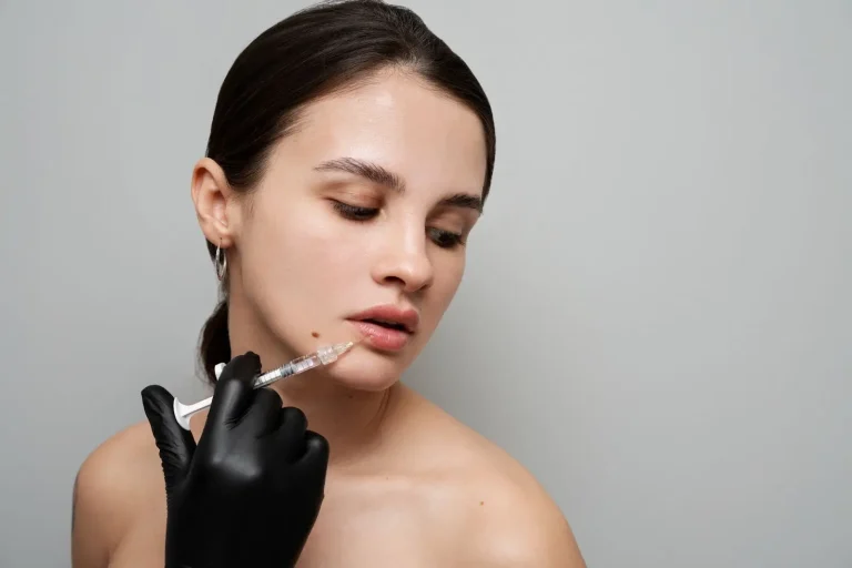 A woman with bare shoulders and dark hair receives an injection in her chin from a person wearing black gloves. She looks downward with a calm expression against a plain light gray background.