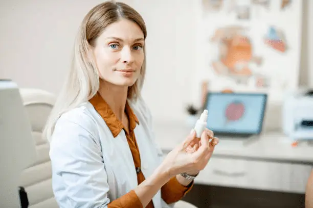 A woman in a white lab coat sits at a desk in a medical office, holding a small white bottle and looking at the camera. A laptop and anatomical posters are visible in the background.