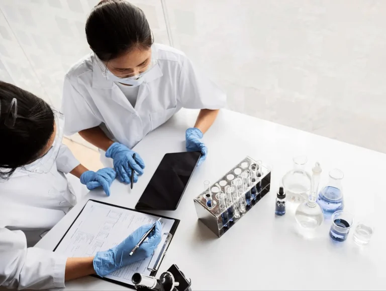 Two scientists in lab coats and gloves work at a white table with a clipboard, tablet, test tubes, and glassware filled with blue liquid, appearing to discuss or analyze their research.