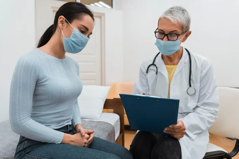 A female doctor in a white coat and face mask holds a clipboard and speaks with a young woman, also wearing a mask, who is sitting with her hands clasped on her lap in a medical office.