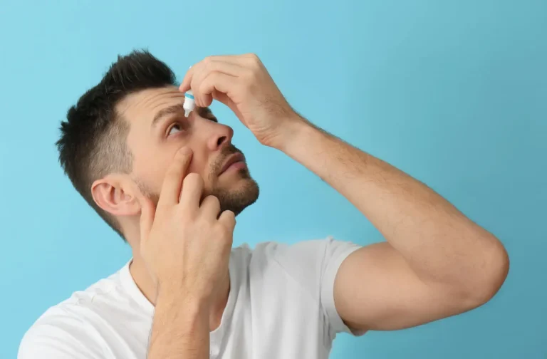A man in a white t-shirt tilts his head back while applying eye drops to his right eye, against a plain blue background.