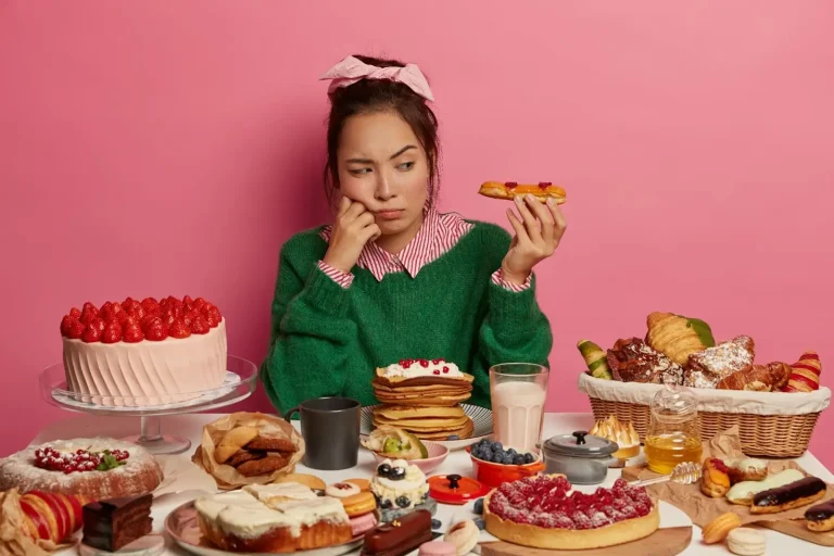 A woman in a green sweater and pink headband sits at a table full of desserts, looking indecisive and holding a pastry. The background is pink and the table is covered with cakes, pancakes, fruit, and pastries.
