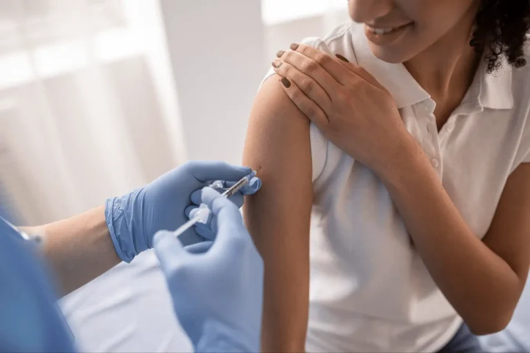 A person wearing a white shirt smiles while a healthcare professional in blue gloves administers a vaccine injection into their upper arm.