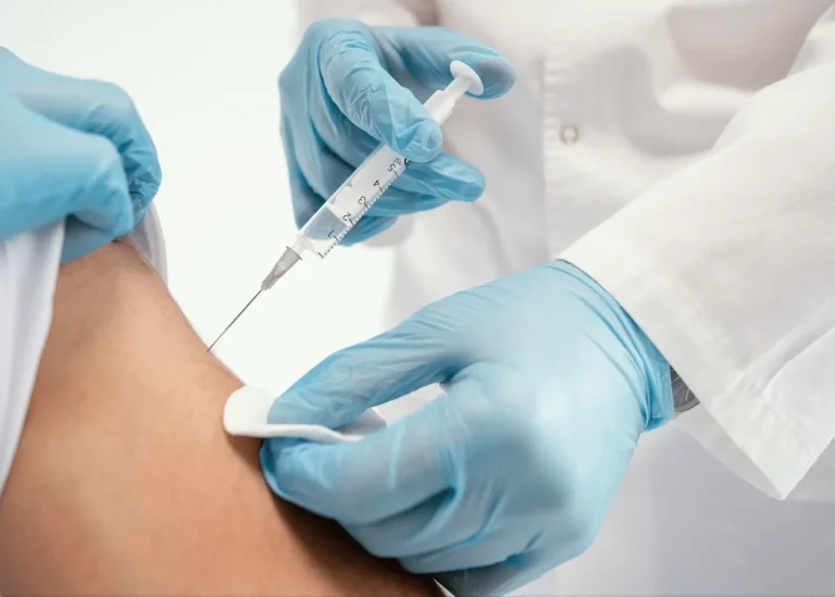 A healthcare worker wearing blue gloves administers an injection with a syringe into a person's upper arm, while holding a cotton pad on the skin.