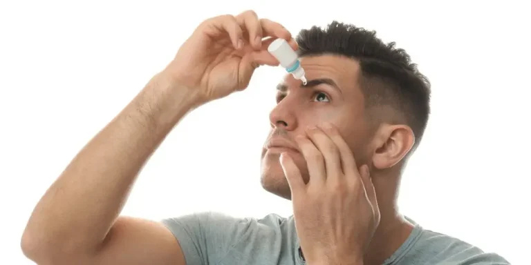 A man tilts his head back and uses one hand to hold open his eye while applying eye drops with the other hand. The background is white.