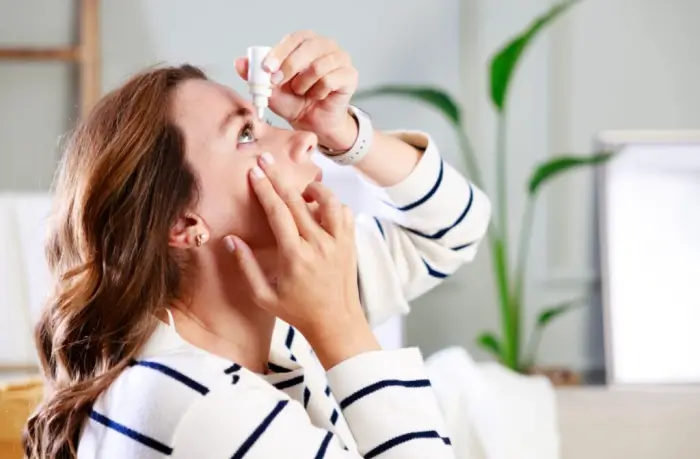 A woman in a striped sweater tilts her head back and uses eyedrops, holding the bottle above her right eye with one hand while pulling down her lower eyelid with the other.