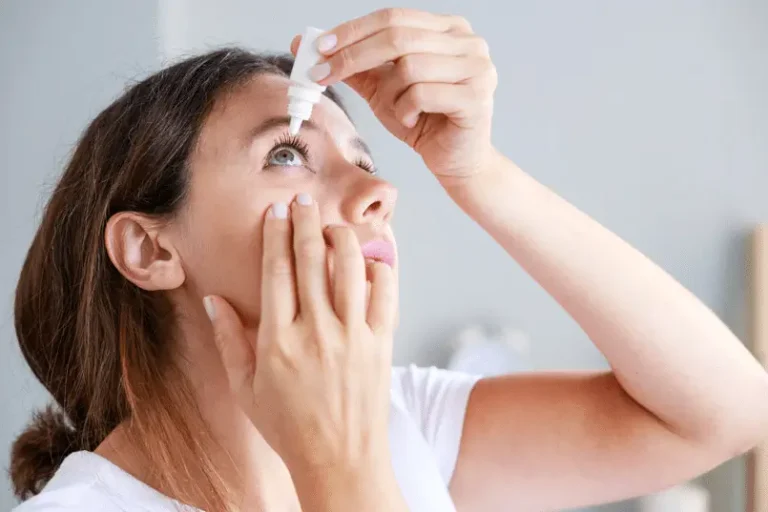 A woman tilts her head back and holds her lower eyelid down while applying eye drops from a small bottle above her eye. She appears to be inside a well-lit room.
