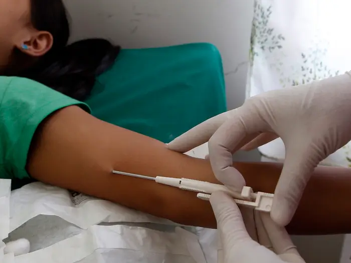 A healthcare worker wearing gloves inserts a needle into a person's arm, likely to draw blood or administer an injection. The person lies on a bed, partially visible.