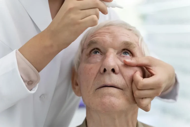 A healthcare professional administers eye drops to an elderly man, gently holding his lower eyelid down as he looks upward.