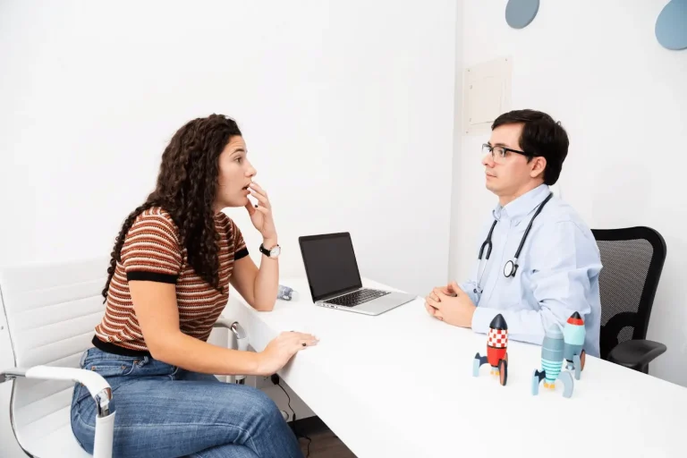 A woman sits across from a doctor at a white desk, appearing concerned as she talks. The doctor listens attentively, wearing a stethoscope. A laptop and toy rockets are on the desk.