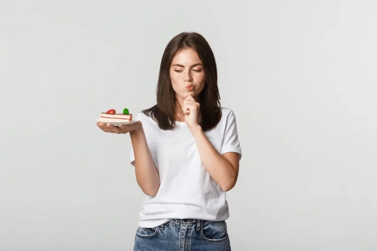A young woman in a white t-shirt and jeans stands holding a slice of cake, looking at it thoughtfully with her lips pursed and one finger resting on her chin, against a plain light background.