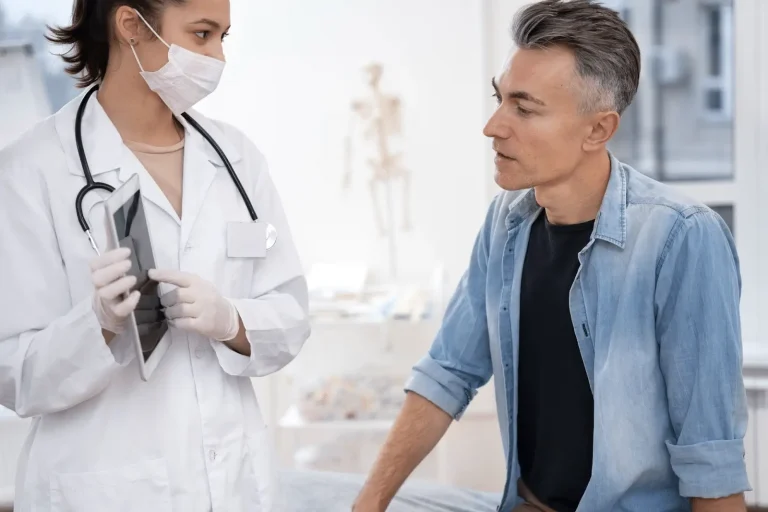 A female doctor wearing a mask and gloves holds a tablet and talks to a seated male patient dressed casually in a denim shirt, inside a bright medical office.
