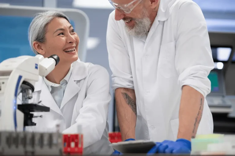 Two scientists in lab coats smile and talk to each other in a laboratory. One stands next to a microscope and red sample tubes, while the other leans on a counter wearing safety glasses and gloves.