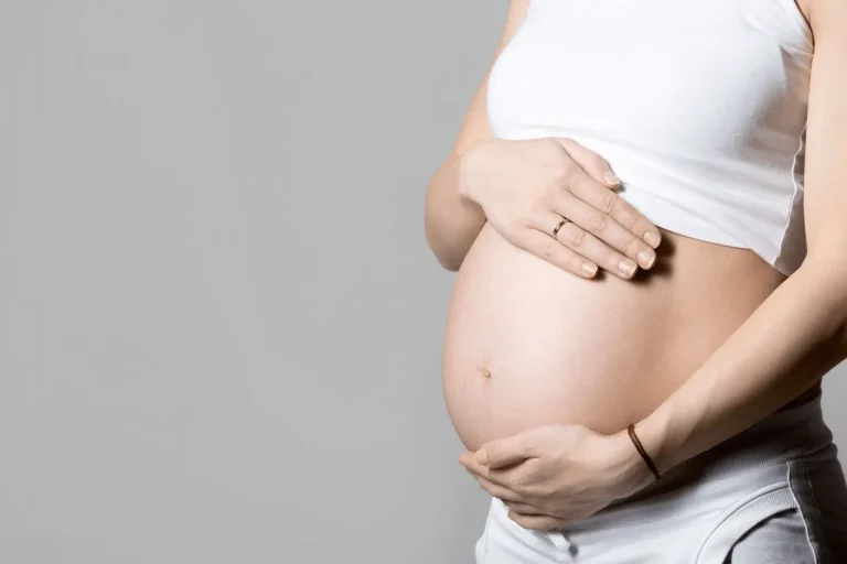 A pregnant person wearing a white top and bottom gently cradles their bare belly with both hands against a plain gray background.
