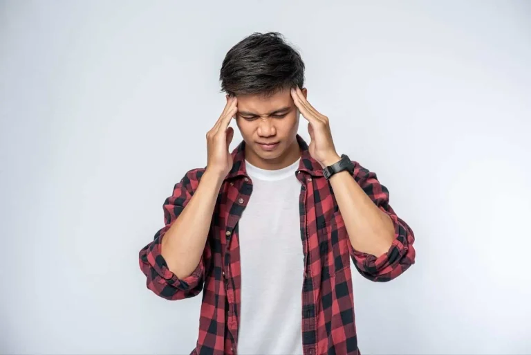 A young person in a red and black plaid shirt stands against a plain background, eyes closed, with both hands pressing their temples, appearing to have a headache or feel stressed.