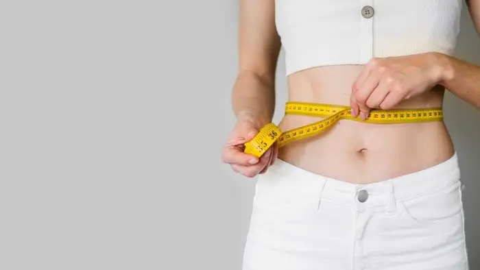 A person wearing a white crop top and white pants measures their waist with a yellow measuring tape, against a plain light gray background.