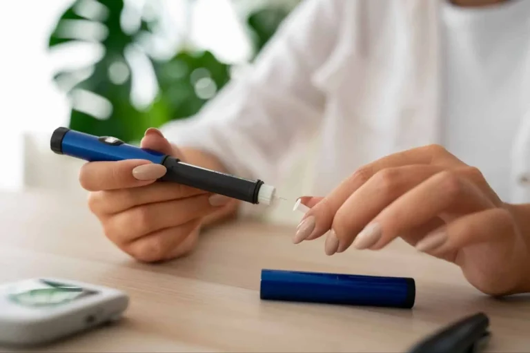 A person holds an insulin pen, preparing to use it, with a glucose meter and pen cap on a table. The background is softly blurred with green plants visible.