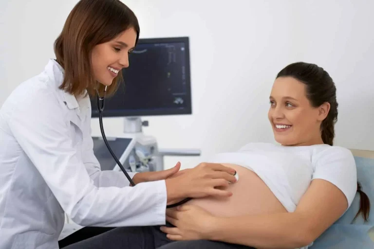 A doctor uses a stethoscope to listen to the belly of a pregnant woman who is lying on an exam table and smiling during a checkup; an ultrasound machine is visible in the background.