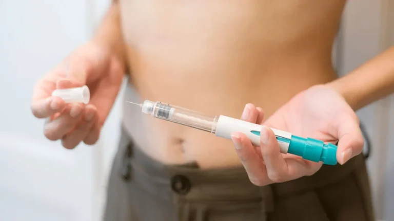 A person holding an insulin pen injector containing the Mounjaro active ingredient in one hand and the cap in the other, standing with their shirt lifted to expose their stomach, preparing for an injection.