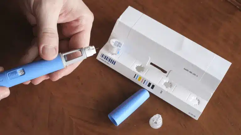 A person is assembling a blue insulin pen next to an open white box with compartments, a blue cap, and a pen needle on a wooden surface.