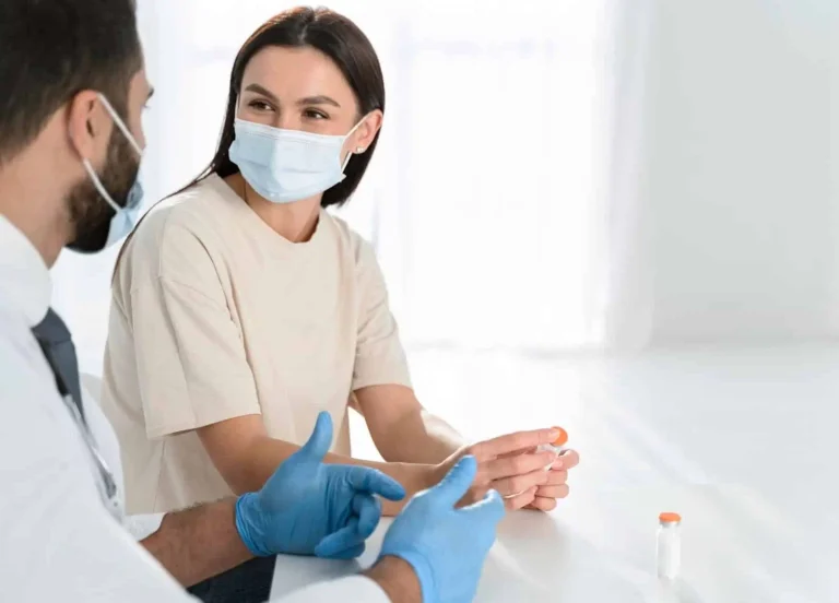 A woman wearing a mask sits at a table with a masked healthcare professional in gloves. The woman holds a vial, and another vial is on the table. They appear to be having a conversation in a bright, clinical setting.