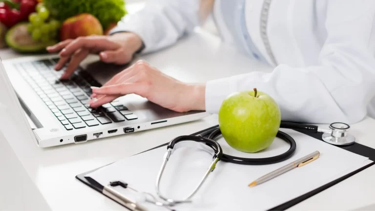 A person in a white coat types on a laptop at a desk with a green apple, stethoscope, pen, and clipboard, suggesting a healthcare or nutrition setting.