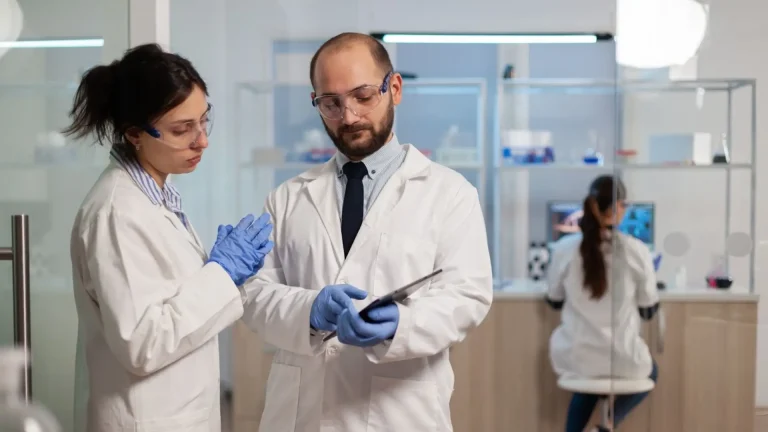 Two scientists in lab coats and safety glasses discuss information on a tablet. One gestures with gloved hands while the other holds the tablet. In the background, another scientist works at a lab bench.