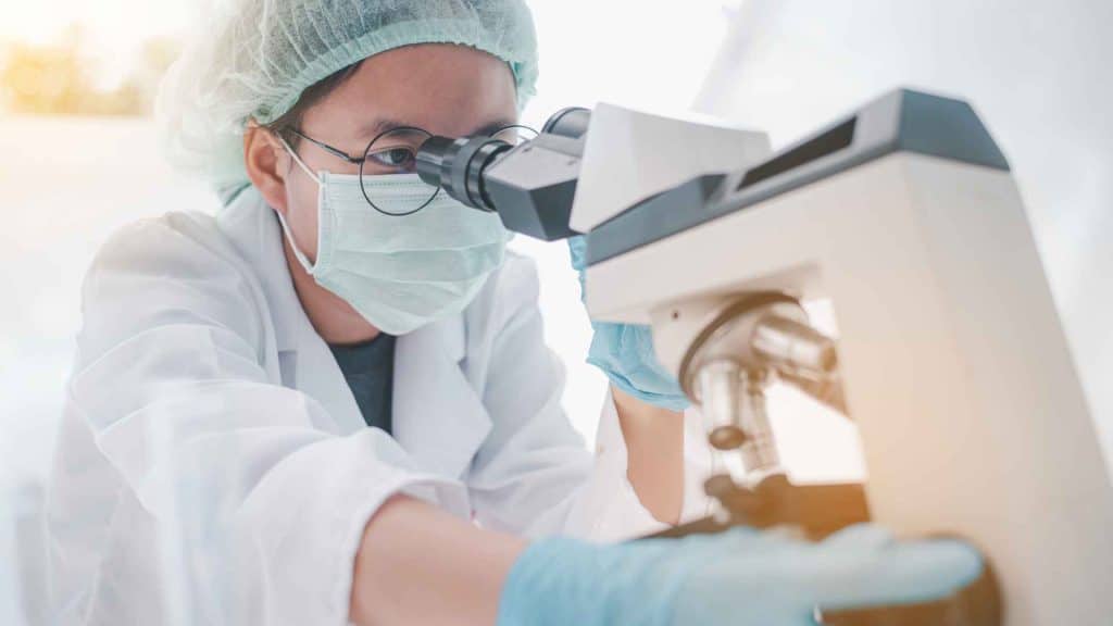 A scientist wearing a lab coat, mask, hairnet, and gloves looks through a microscope in a laboratory setting.