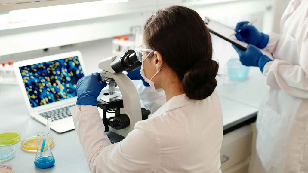 A scientist looks into a microscope in a laboratory, wearing gloves and a lab coat. Another person holds a clipboard.