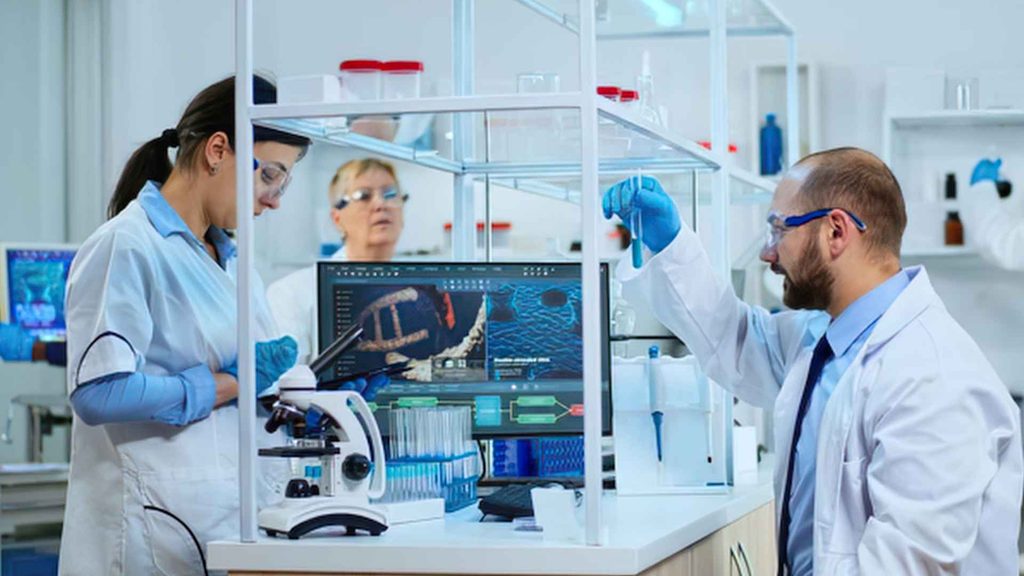 Three scientists in lab coats and safety glasses work in a laboratory with computers, test tubes, and a microscope visible on the counter.