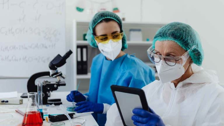 Two scientists wearing masks, protective gowns, and hairnets work in a laboratory, with one person examining a tablet while the other works with laboratory equipment.