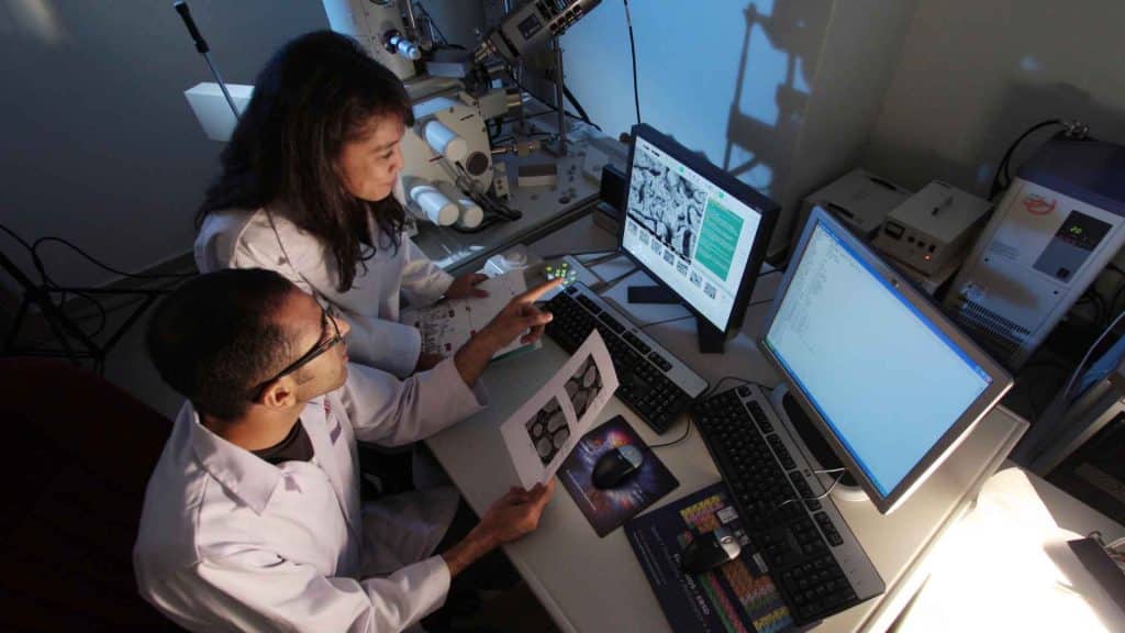 Two researchers in lab coats reviewing digital images and printed data at a computer workstation.