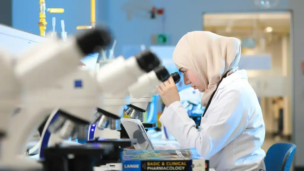 A researcher wearing a hijab and white lab coat examines a sample through a microscope in a laboratory.