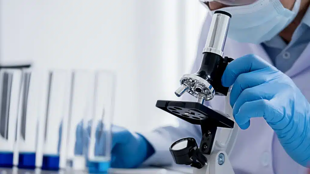 A scientist in a lab coat, mask, and blue gloves adjusts a microscope while test tubes sit on the table nearby.
