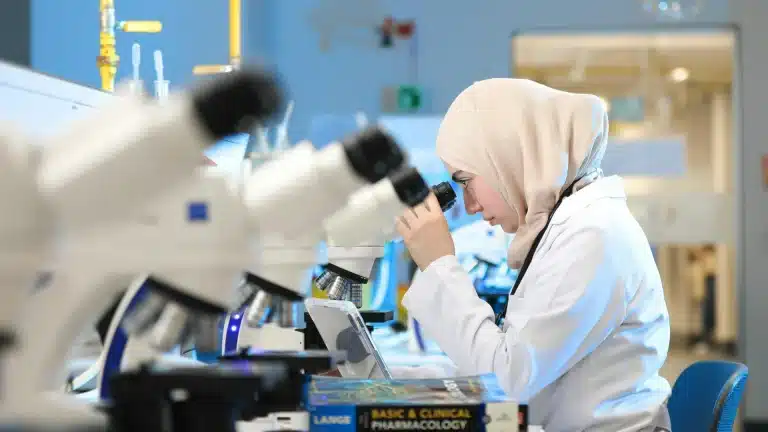 A researcher wearing a hijab and white lab coat examines a sample through a microscope in a laboratory.